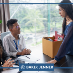 A woman stands holding a box of personal items while two people sit across a desk from her, appearing to speak with her in an office setting. The scene is branded with the Baker Jenner logo.